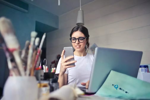 Une femme à lunettes regarde son smartphone devant un ordinateur portable, des pinceaux au premier plan