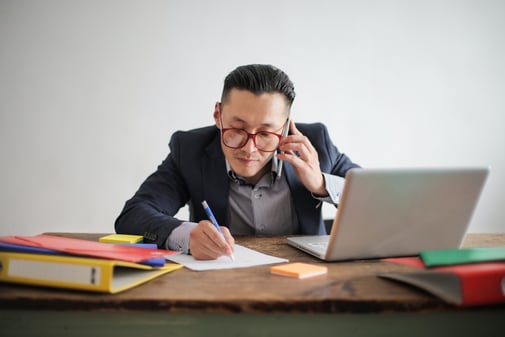Un homme au téléphone prend des notes à son bureau, entre un ordinateur portable et des classeurs