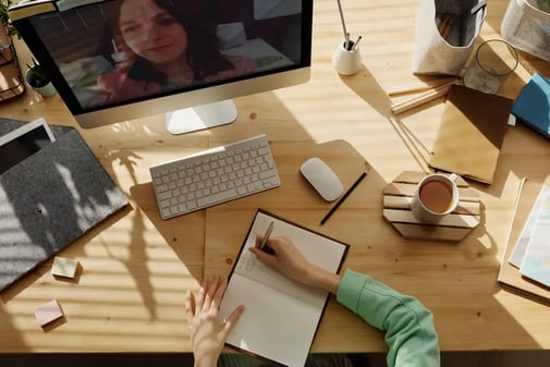 Vue du dessus d'un bureau en bois : une main prend des notes dans un carnet face à une visioconférence sur l'écran