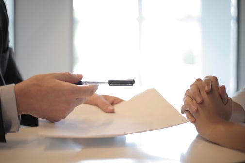 Une personne tend un stylo et un document à compléter à son interlocuteur, mains jointes sur la table