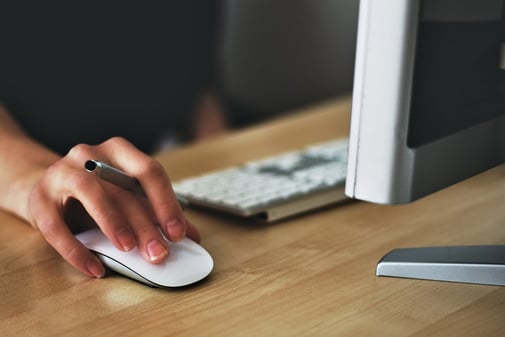Une main sur une souris blanche, un stylo entre les doigts, devant un clavier et un écran sur un bureau en bois